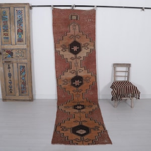 May include: A long, narrow runner rug featuring a terracotta and tan background with a repeating geometric pattern of dark brown hexagonal medallions. The rug displays symmetrical diamond-shaped motifs and small star-like accents throughout the design.