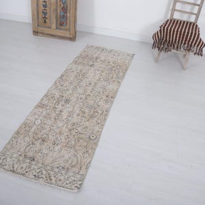 May include: A long, rectangular runner rug featuring a faded, neutral-toned pattern with intricate floral and geometric motifs in shades of beige, cream, and soft gray. The rug has a distressed texture and a decorative border along the short edges.