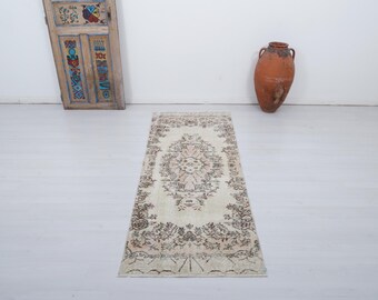 May include: A rectangular, vintage-style rug with an intricate floral pattern in cream, brown, and beige tones. The rug is placed on a white floor, with a decorative wooden door and a large terracotta vase in the background.