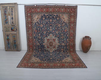 May include: A large, ornate rug with a navy blue field and a detailed floral pattern in shades of red, beige, and cream. The rug is displayed against a white wall, with a decorative wooden door and a terracotta vase nearby.