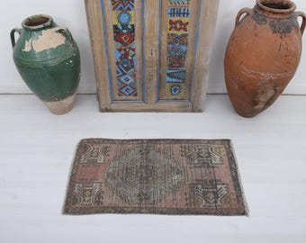 May include: A small, rectangular rug with a faded, distressed appearance. The rug features a central design in shades of gray and brown, framed by a pink border. The rug is placed on a white surface, with a green vase, a wooden panel, and a brown vase in the background.