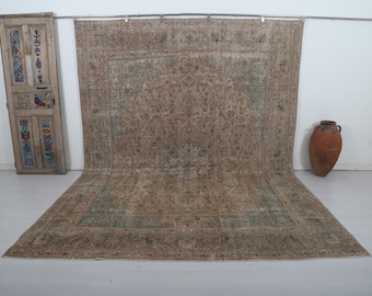 May include: A large, rectangular, distressed-look rug with a central floral pattern in shades of beige, brown, and teal. The rug is displayed against a white wall, with a decorative wooden door and a terracotta vase nearby.