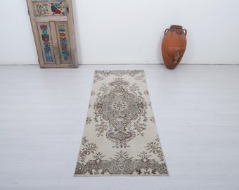 May include: A rectangular, cream-colored rug with an intricate, symmetrical floral pattern in shades of brown and gray. The rug is placed on a white floor, with a decorative wooden door and a large terracotta vase in the background.