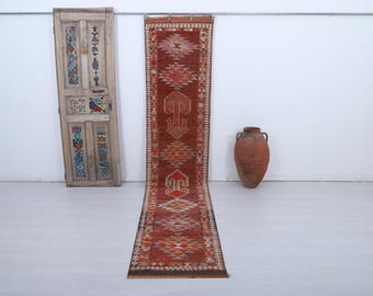 May include: A long, rectangular, vintage rug with a geometric pattern in shades of red, orange, and beige. The rug is displayed on a white floor, with a decorative wooden door and a large terracotta vase in the background.