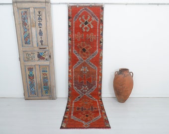 May include: A long, narrow, red and orange rug with a geometric pattern. The rug has a dark brown border and is hanging on a white wall.