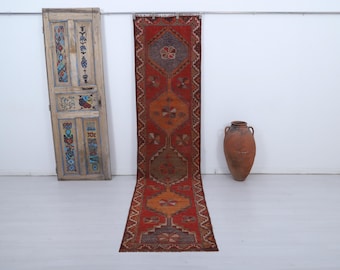 May include: A long, rectangular, red and orange patterned rug with a geometric design. The rug is displayed on a white floor, with a decorative wooden door and a large brown vase in the background.