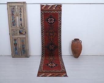 May include: A long, rectangular red rug with geometric patterns in black, brown, and white. The rug is displayed on a white floor against a white wall. An antique wooden door and a large terracotta vase are also in the image.