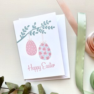 May include: A white card with a green branch with two pink Easter eggs hanging from it. The eggs are decorated with pink and white floral patterns. The text "Happy Easter" is written in pink below the eggs.