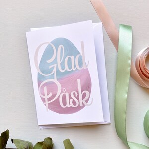 May include: A white card with a pink and blue watercolor egg design. The text "Glad Påsk" is written in white on the egg.