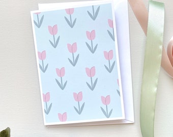 May include: A greeting card with a light blue background featuring a pattern of pink tulip illustrations with gray stems and leaves. The card is inside a white envelope. Ribbons in shades of pink and green are in the background.