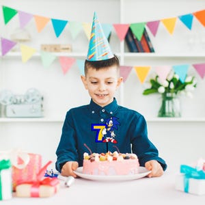 May include: A young boy wearing a blue shirt with a Sonic the Hedgehog graphic and the number 7 on it, is celebrating his birthday with a pink cake decorated with fruit and a blue and white party hat on his head.