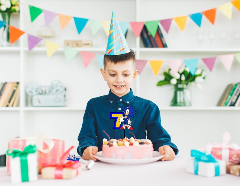 May include: A young boy wearing a blue shirt with a Sonic the Hedgehog graphic and the number 7 on it, is celebrating his birthday with a pink cake decorated with fruit and a blue and white party hat on his head.