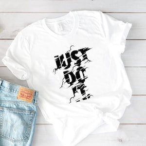 May include: White t-shirt with the words "JUST DO IT" in a black, stylized font. The text has a cloud-like design around it. The shirt is folded and placed on a light wood surface next to a pair of blue jeans.