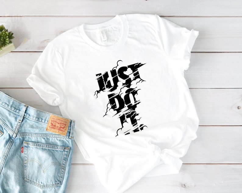 May include: White t-shirt with the words "JUST DO IT" in a black, stylized font. The text has a cloud-like design around it. The shirt is folded and placed on a light wood surface next to a pair of blue jeans.
