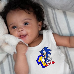 May include: A baby wearing a white sleeveless onesie with a blue and yellow graphic of Sonic the Hedgehog and the number one. The baby is lying on a striped blanket.