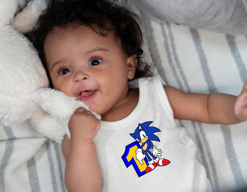 May include: A baby wearing a white sleeveless onesie with a blue and yellow graphic of Sonic the Hedgehog and the number one. The baby is lying on a striped blanket.