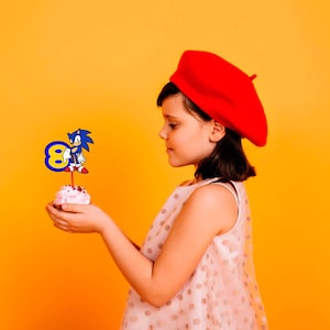 May include: A young person wearing a red beret holds a cupcake with a blue and yellow Sonic the Hedgehog character topper. The cupcake is on a yellow background.
