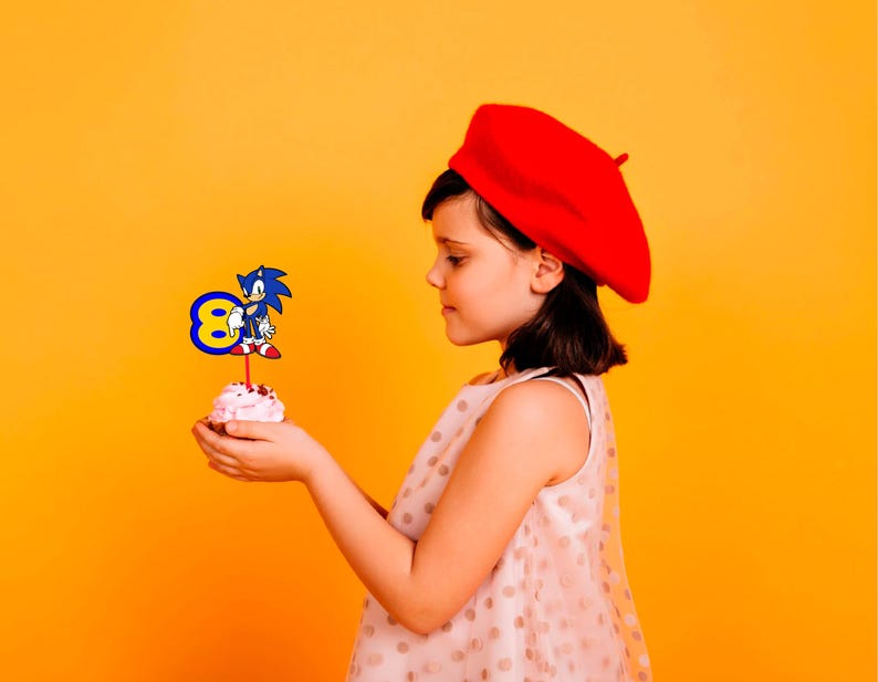 May include: A young person wearing a red beret holds a cupcake with a blue and yellow Sonic the Hedgehog character topper. The cupcake is on a yellow background.