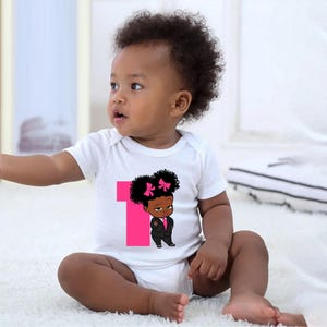 May include: A white baby onesie featuring a pink number one and a cartoon image of a little girl with a pink bow in her hair. The girl is wearing a black suit and has a serious expression on her face.