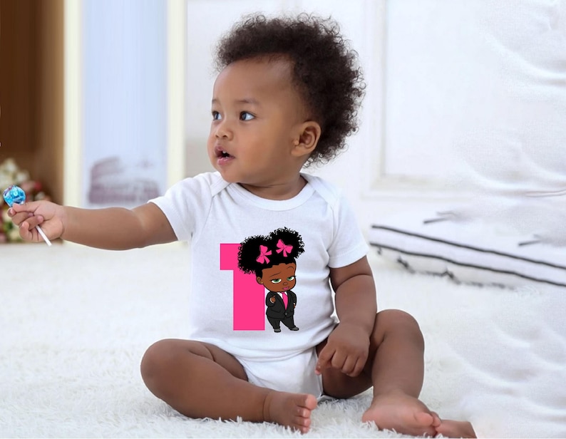 May include: A white baby onesie featuring a pink number one and a cartoon image of a little girl with a pink bow in her hair. The girl is wearing a black suit and has a serious expression on her face.
