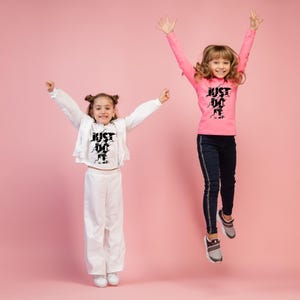 May include: Two young people jumping in the air, wearing athletic clothing. One wears a pink long-sleeve shirt and dark blue pants. The other wears a white outfit. Both outfits have the text "JUST DO IT" in black.
