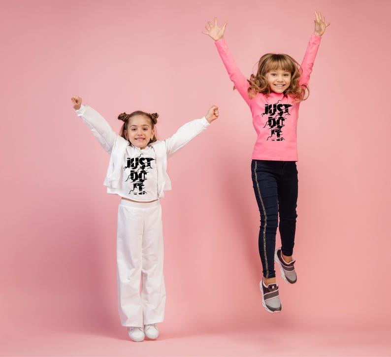 May include: Two young people jumping in the air, wearing athletic clothing. One wears a pink long-sleeve shirt and dark blue pants. The other wears a white outfit. Both outfits have the text "JUST DO IT" in black.
