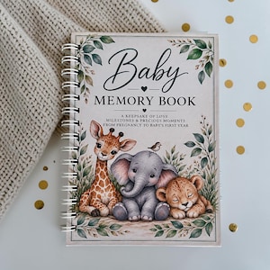 May include: A spiral-bound baby memory book with a cream-coloured cover featuring illustrations of a giraffe, an elephant, and a lion cub surrounded by green leaves. The text reads Baby MEMORY BOOK A KEEPSAKE OF LOVE MILESTONES & PRECIOUS MOMENTS.