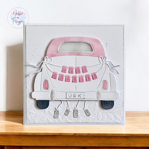 May include: A white and pink wedding card featuring a car with a banner that reads "Just Married." The car has a pink roof and accents, with the initials "J & K" on the license plate. Decorative elements include silver charms and floral embossing.