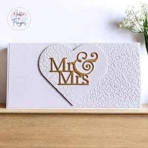 May include: A white card with a gold heart-shaped design featuring the words "Mr & Mrs" in a decorative font. The card has a white embossed floral pattern.