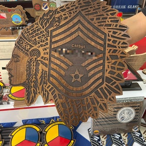 May include: A wooden plaque featuring a Native American profile and military insignia. The plaque is brown with intricate laser-cut details, including a star and the text "CMSgt". The design incorporates feathers and geometric patterns, with other military emblems in the background.