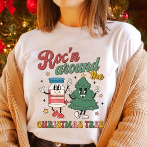 May include: White t-shirt with a retro-style graphic of a cartoon Christmas tree and a vial labeled "Rocuronium 100 mg per 10 ml" with the text "Roc'n around the Christmas Tree".
