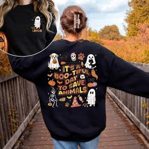 Può includere: Felpa nera con grafica a tema Halloween. Il design include fantasmi, zucche, un cane in costume da fantasma e la scritta "It's a Boo-tiful Day to Save Animals".