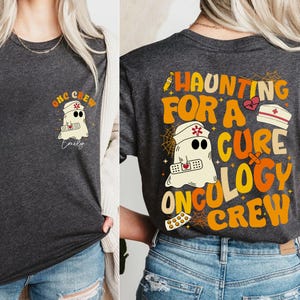 Può includere: Una maglietta grigia scura con un fantasma che indossa un cappello da infermiera e tiene un bendaggio. Il testo sulla maglietta recita "Haunting for a Cure Oncology Crew".