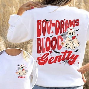 Könnte beinhalten: Weißes Sweatshirt mit dem roten Text "Boo-Draws Blood Gently" und einem Cartoon-Geist mit Krankenschwestermütze und Gesichtsmaske. Das Design umfasst Fledermäuse, Spinnweben und Sterne, ideal für Halloween oder medizinisches Personal.
