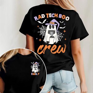 Op de afbeelding: Zwart T-shirt met een Halloween-thema ontwerp. De achterkant heeft een spook met een heksenhoed, met de tekst "RAD TECH BOO crew". Een kleinere versie van het ontwerp staat op de voorkant, met de naam "EMILY".