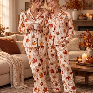Puede incluir: Dos conjuntos a juego de camisas de pijama de manga larga con botones y pantalones largos de pijama en tela de satén color crema. Las prendas presentan un estampado otoñal repetitivo de hojas de arce naranjas y rojas, bellotas y calabazas.
