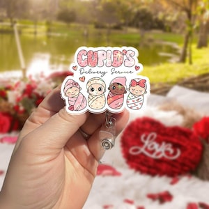 Op de afbeelding: Een wit en roze badge reel met de tekst "Cupid's Delivery Service" en illustraties van vier baby's. De baby's zijn in dekens gewikkeld en dragen hoeden. De badge reel wordt in een hand gehouden.