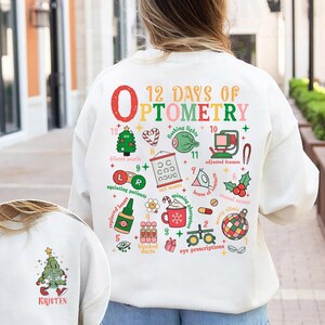 May include: A white sweatshirt with a colorful graphic design featuring the text "12 Days of Optometry" and various optometry-related illustrations. The design includes a Christmas tree, eye charts, and other festive elements. The name "Kristen" is printed on the lower left.