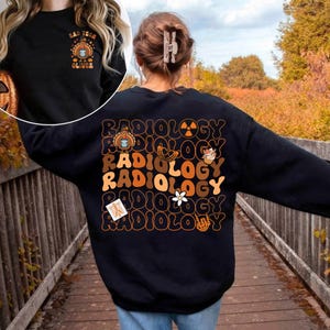 Puede incluir: Sudadera negra con la palabra "RADIOLOGY" en fuente retro naranja y marrón. La parte trasera de la sudadera tiene varios gráficos, incluyendo un pavo, un plátano, una magdalena, una flor y una radiografía. La parte delantera de la sudadera tiene el texto "BAD TECH OLIVER".