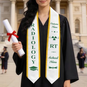 May include: A graduation sash with the word "RADIOLOGY" in green lettering on a white background with a gold border. The sash also features a graduation cap graphic and the text "CLASS OF 2023". The person is wearing a black graduation gown and holding a diploma.