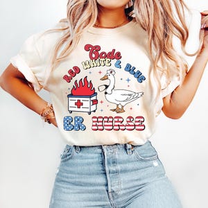 May include: Cream-colored t-shirt with a graphic design featuring a goose wearing a nurse's hat, a medical box with flames, and the text "Code White & Blue ER Nurse" in red, white, and blue.
