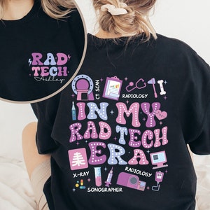 Op de afbeelding: Zwart sweatshirt met een roze en paarse grafische print met de tekst "In My Rad Tech Era". De print bevat verschillende iconen van medische apparatuur, zoals een röntgenfoto, een stethoscoop en een CT-scanner.