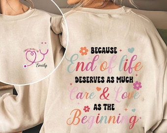 May include: A beige sweatshirt with a pink stethoscope and the text "Hospice Nurse" and "Emily" on the left side. The right side of the sweatshirt has the text "Because End of Life Deserves As Much Care & Love As The Beginning" in colorful letters.
