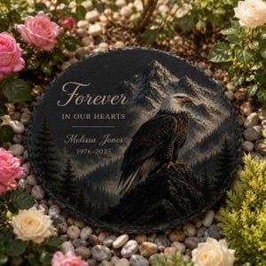 May include: A round black slate memorial plaque featuring a bald eagle perched on a rocky mountain peak. The text reads Forever IN OUR HEARTS, Melissa Jones, and 1976-2025. The design includes a detailed landscape of pine trees and mountains.