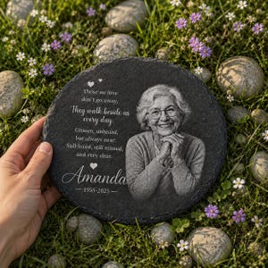 May include: A round black slate memorial plaque featuring an engraved portrait of an older person with glasses and short hair. The text reads: Those we love don't go away, They walk beside us every day. Unseen, unheard, but always near, Still loved, still missed, and very dear. Amanda 1955-2025.