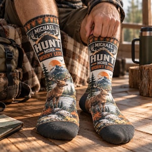 May include: A pair of crew socks featuring a printed outdoor scene with a deer, mountains, and a sunset. The text reads MICHAEL HUNT MORE WORRY LESS with a rifle graphic. The socks have dark gray toes and heels with a forest pattern at the top.