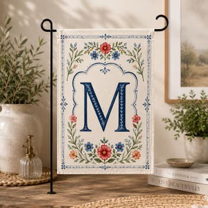 May include: A cream-colored garden flag with a navy blue letter "M" in the center, surrounded by a floral wreath of red and blue flowers. The flag is displayed on a black metal stand, perfect for outdoor decor.