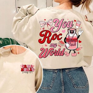 Może przedstawiać: Beżowa bluza z różowo-czerwonym nadrukiem graficznym z napisem "You Roc my World" i postacią kreskówkowego flakonu medycznego. Na plecach bluzy znajduje się różowo-czerwony nadruk graficzny z napisem "CRNA NURSE Emily. RVT".