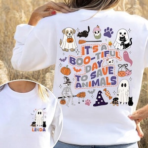 Puede incluir: Sudadera blanca con un diseño de Halloween. El diseño incluye un fantasma, un perro, un gato, una calabaza y el texto "It's a Boo-tiful Day to Save Animals". Una versión más pequeña del diseño está en la parte delantera.