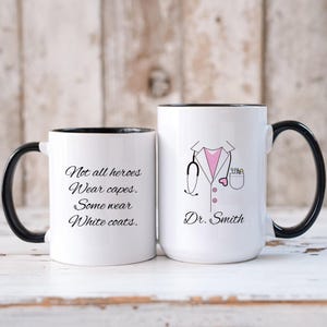 May include: Two white coffee mugs with black rims. One mug has a black handle and says "Not all heroes wear capes. Some wear white coats." The other mug has a black handle and a black and white illustration of a doctor's coat with a stethoscope, a pink shirt, and a heart in the pocket. The text "Dr. Smith" is below the illustration.